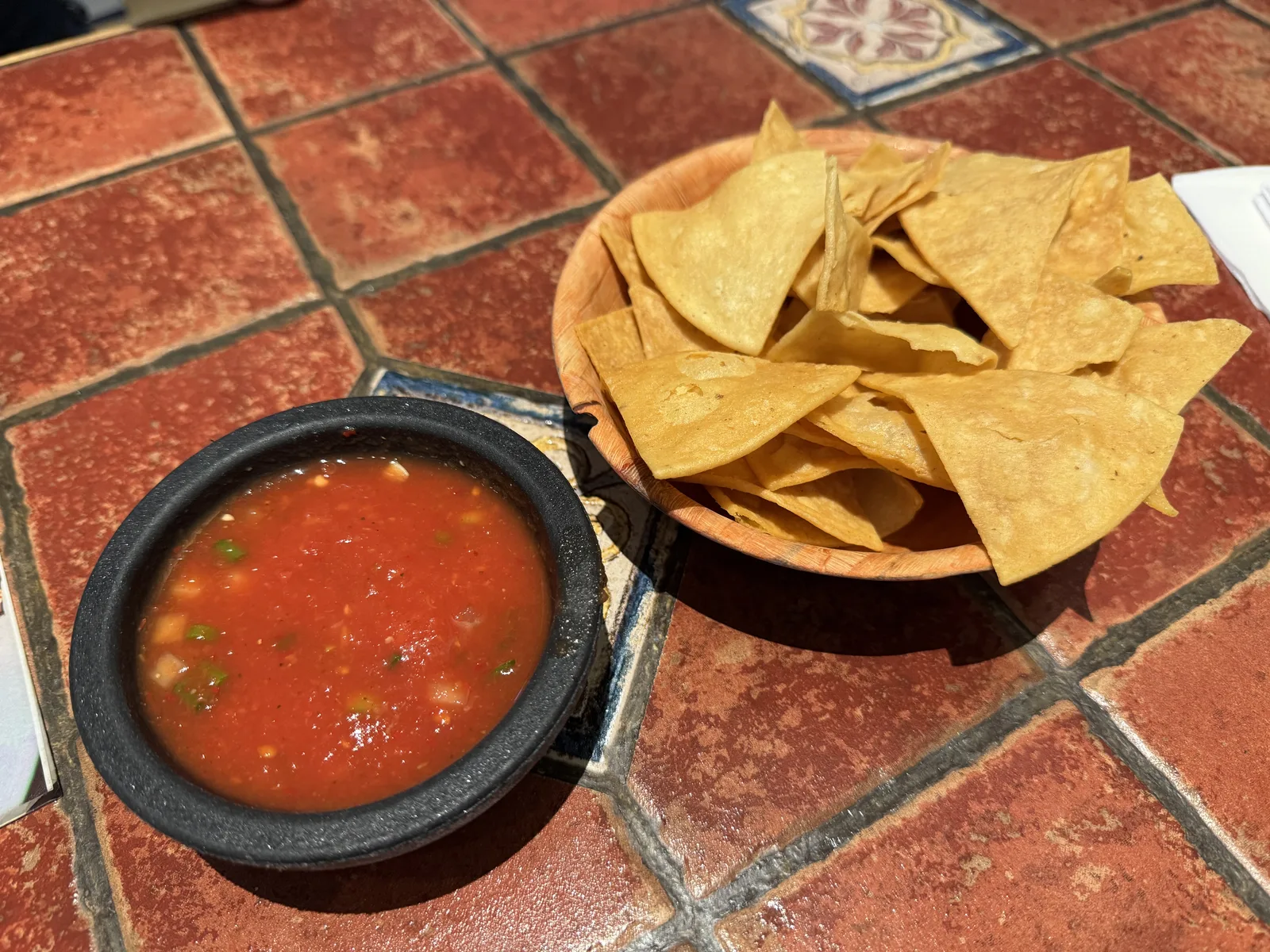 A stone bowl of red salsa beside a bowl of tortilla chips on a tiled table
