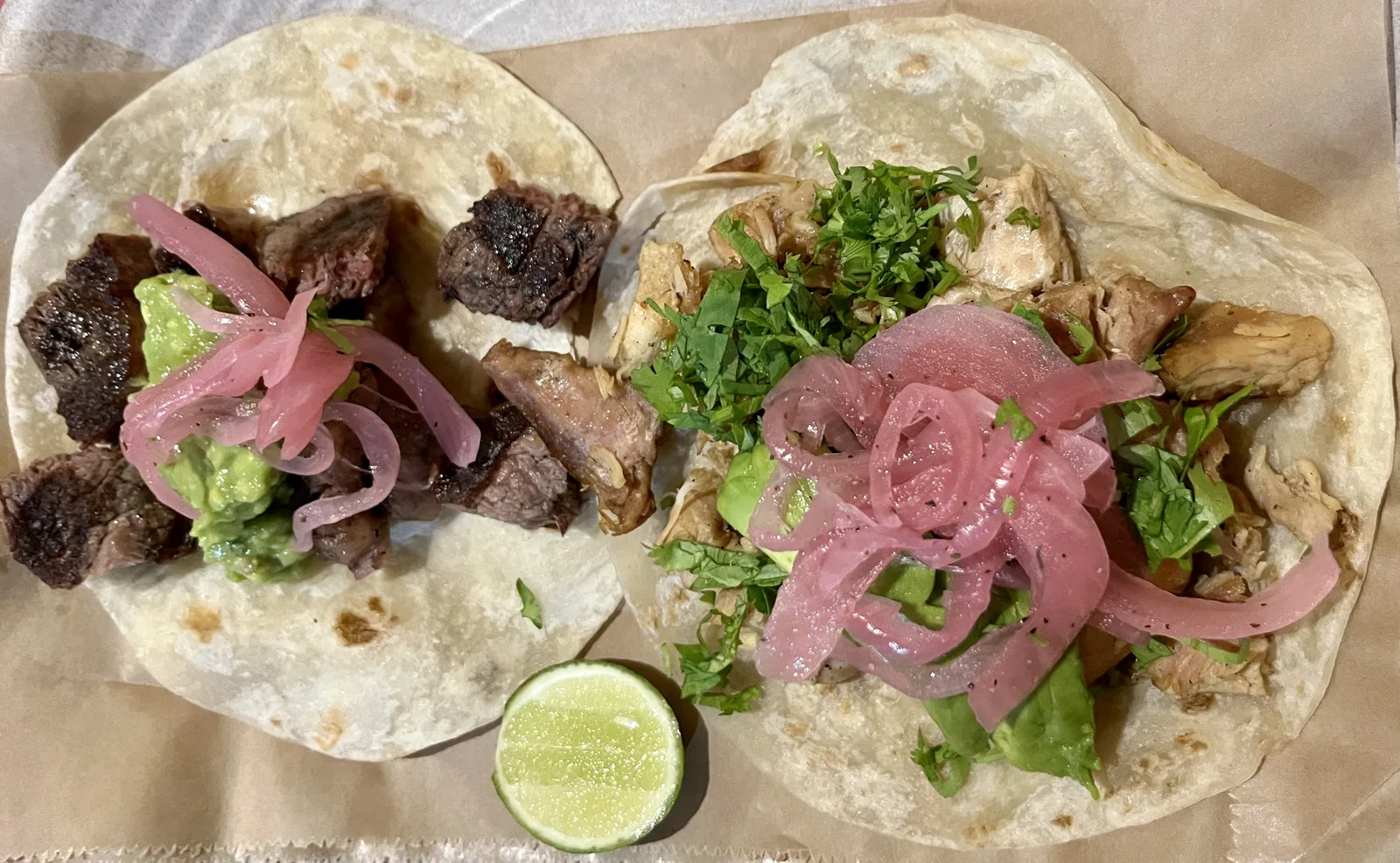 Two tacos with grilled steak and chicken, pickled red onion and cilantro, beside half a lime